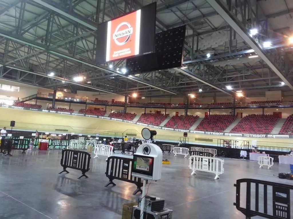 Borne photo au bord de la piste d'un vélodrome, écran suspendu au-dessus des gradins