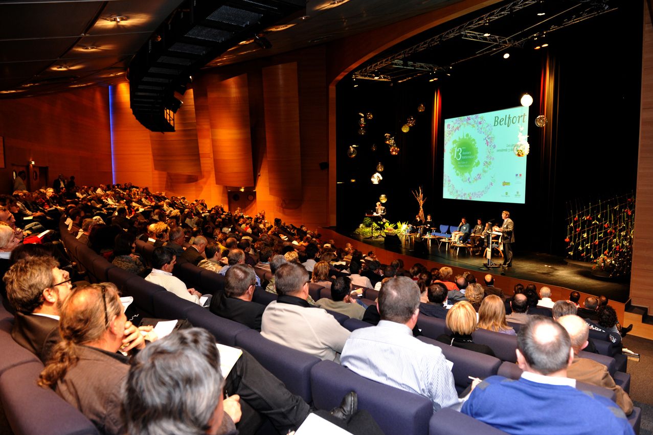 Auditorium plein face à une scène et un grand écran, public de dos