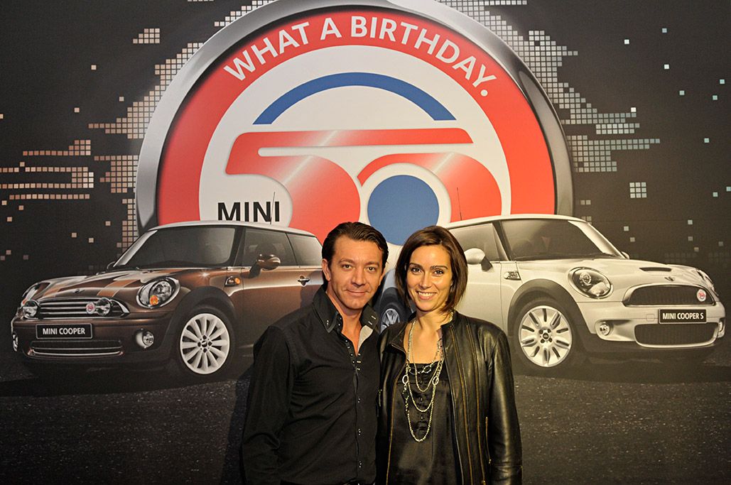 Couple posant devant le photocall des 50 ans de la MINI, deux voitures sur le visuel