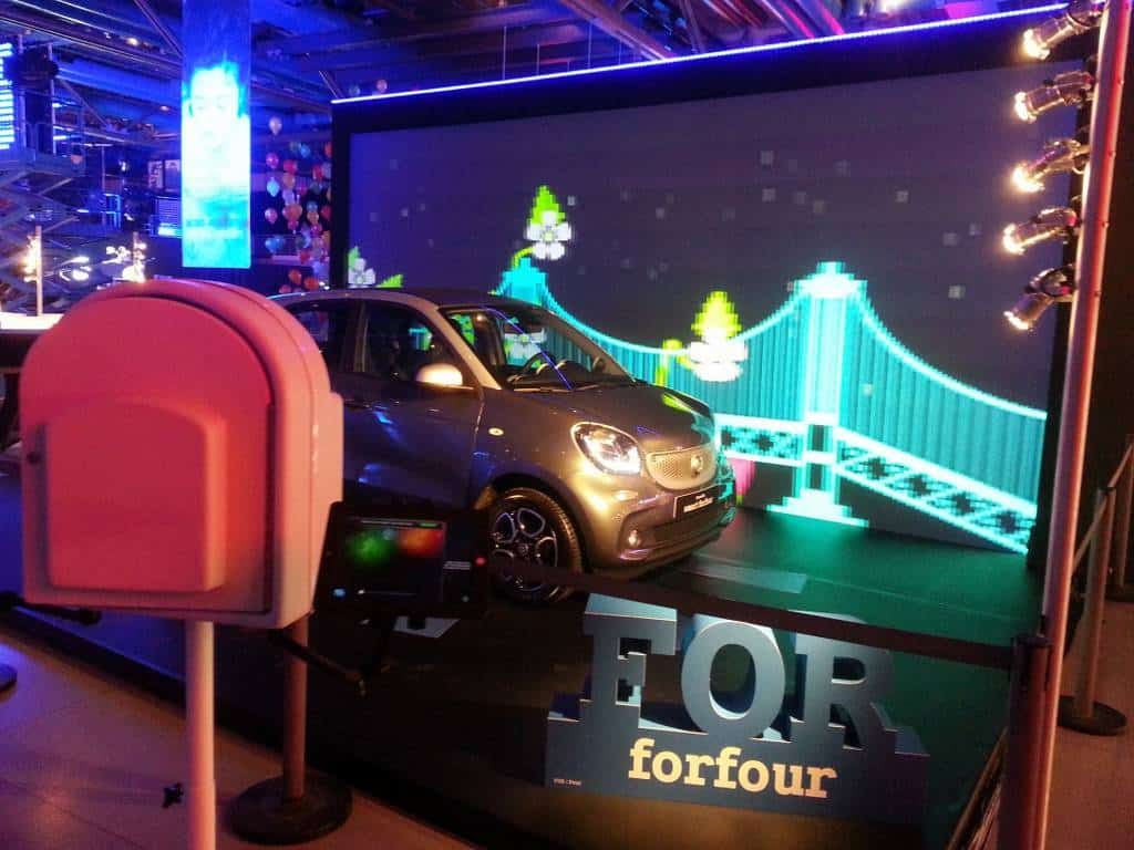 Borne photo rouge devant une smart forfour et un pont lumineux sur écran géant