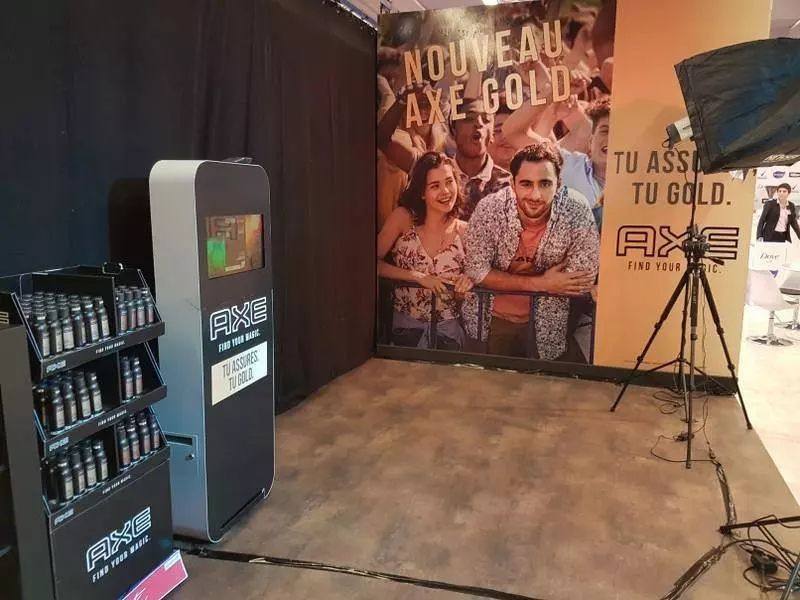 Borne photo Axe à côté de l'affiche du parfum Axe Gold et d'un présentoir de produits