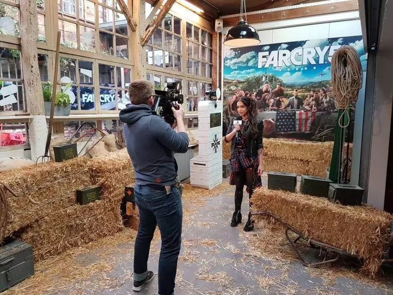 Cameraman interviewant une invitée dans le décor Far Cry 5, borne photo en arrière-plan