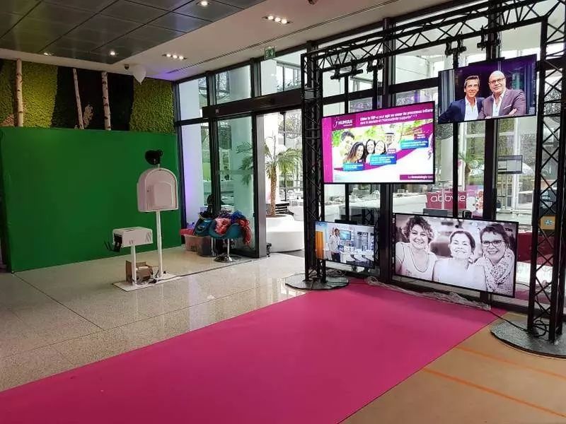Fond vert et borne à l'entrée d'un salon, tapis rose et structure d'écrans