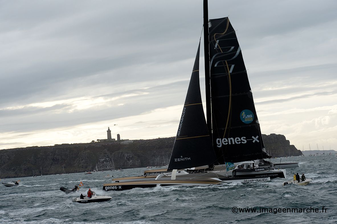 Trimaran géant Spindrift 2 aux voiles noires passant devant la pointe et son phare